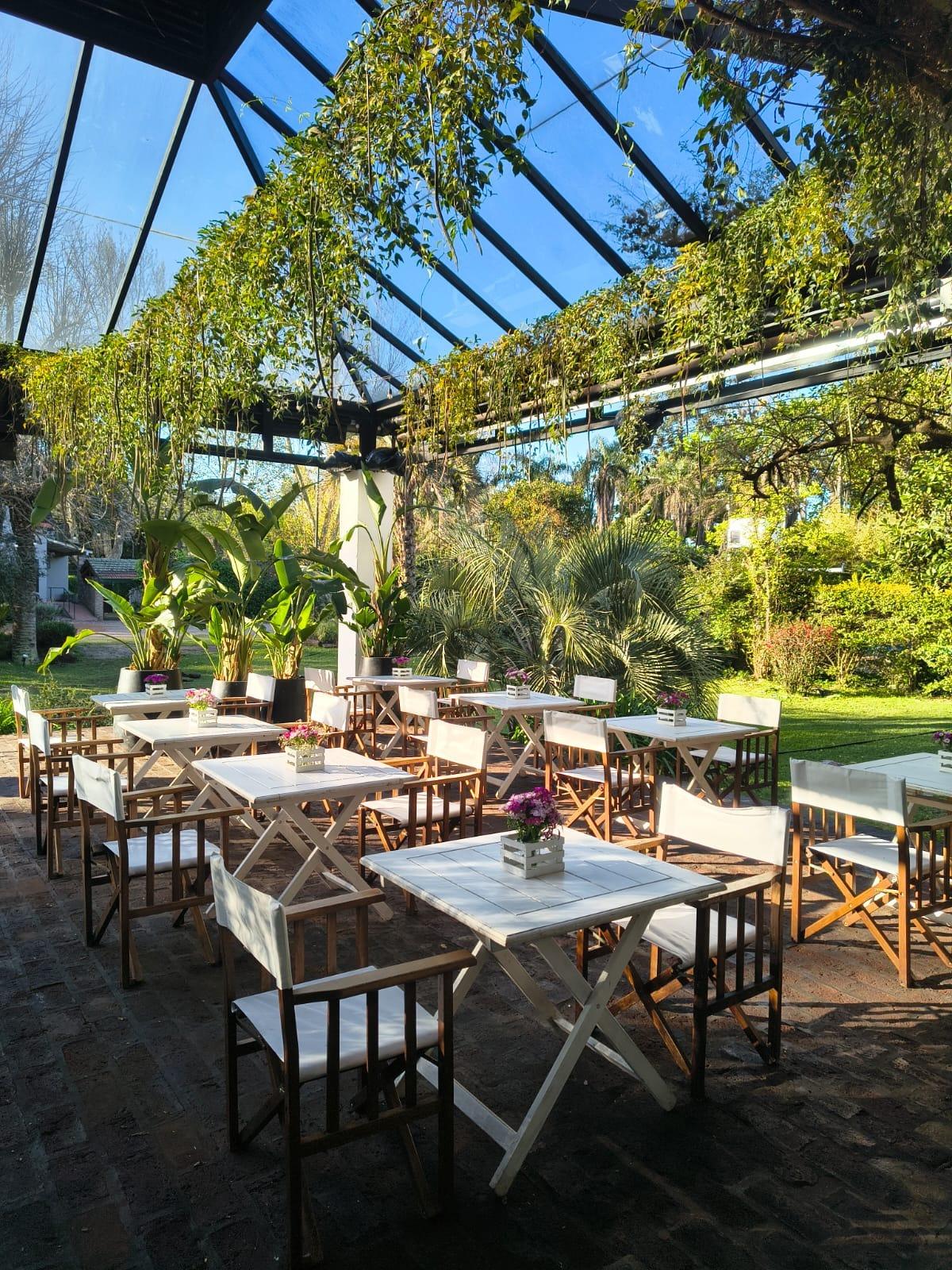 Pérgola vidriada al sol, con mesas blancas y el parque alrededor